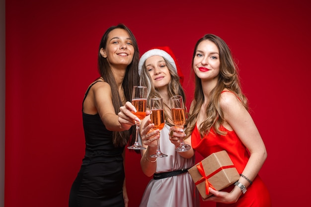 Amigos felices de las mujeres jóvenes con regalo y champán celebrando las vacaciones juntos en fiesta en la pared roja, espacio de copia