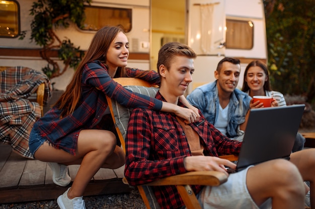 Amigos felices usando laptop cerca de rv de picnic en el camping en el bosque. Los jóvenes tienen una aventura de verano en autocaravana, autocaravana Ocios para dos parejas, viajando con remolque