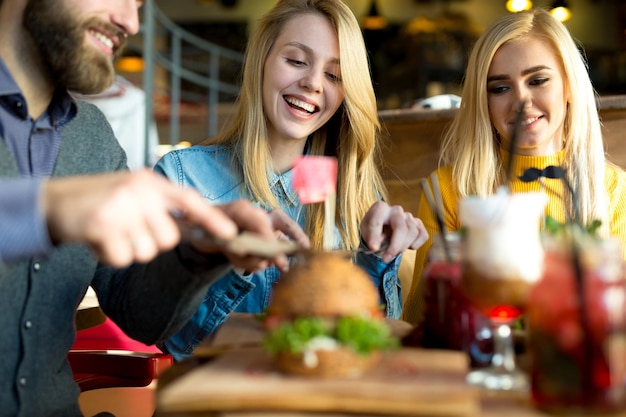 Foto amigos felizes cortam um hambúrguer com uma faca em um restaurante e riem.