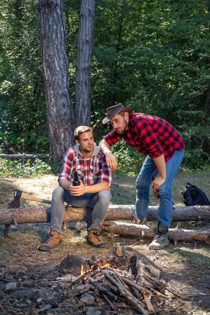 Amigos felizes em uma viagem de acampamento relaxando pela fogueira Grupo de amigos desfrutando de piquenique na floresta e bebendo cerveja Dois lenhadores fazendo piquenique Turistas relaxando