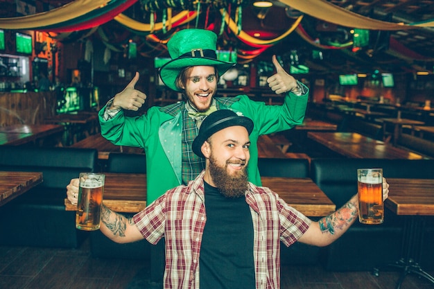 Amigos felizes estão no pub e pose. Eles parecem retos. Cara de terno verde aparecer seus grandes polegares. Jovem na frente tem duas canecas de cerveja nas mãos.