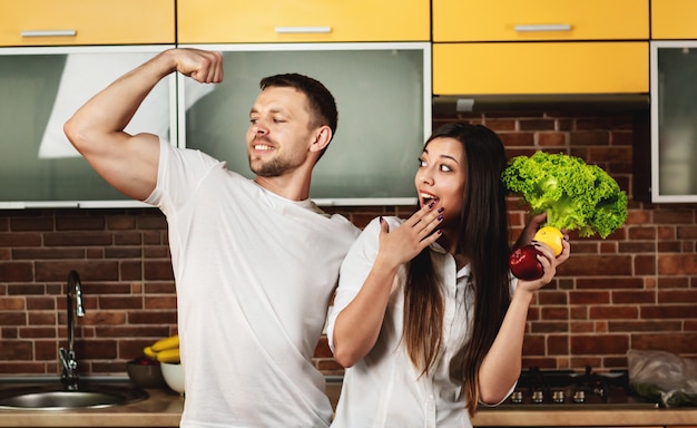 Amigos felizes preparando comida para o jantar, posando na cozinha segurando frutas e legumes. homem mostra bíceps no braço. promovendo uma alimentação saudável