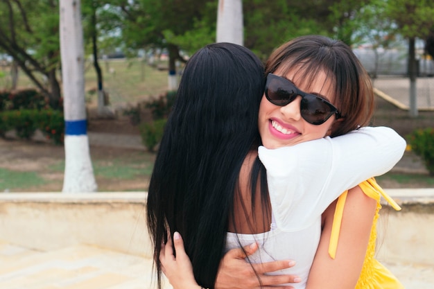 Amigos femininos latinos, abraçando-se juntos de pé em um parque.