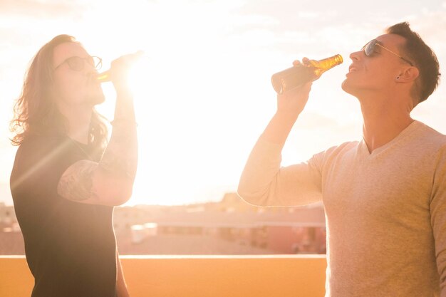 Foto amigos hombres bebiendo cerveza mientras están de pie contra el cielo durante la puesta de sol