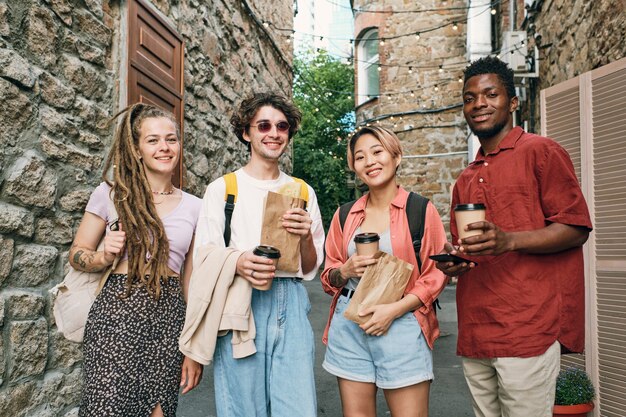 Amigos interculturales felices con bebidas y bocadillos de pie entre edificios de piedra