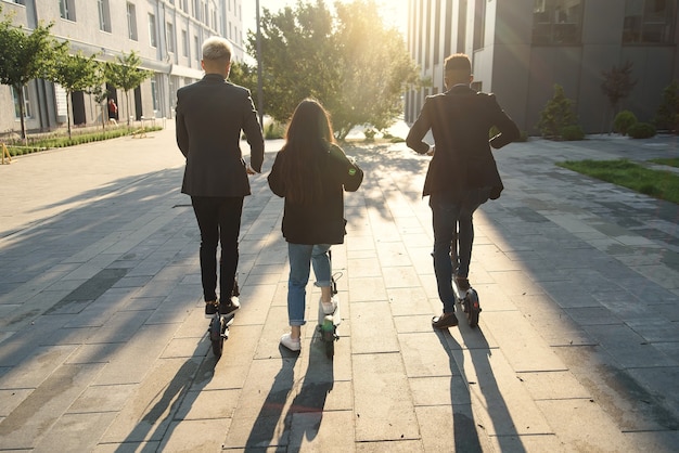 Amigos internacionais alegres segurando scooters elétricos perto de um prédio moderno