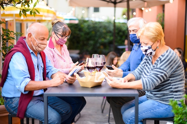 Amigos jubilados con mascarilla viendo el teléfono inteligente mientras beben vino tinto en el restaurante