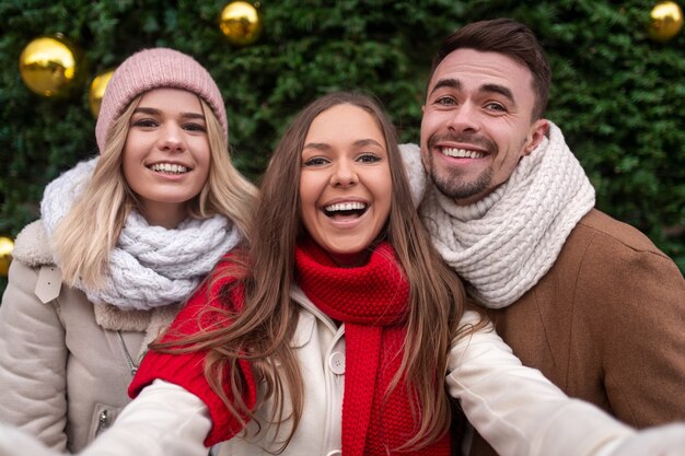 Amigos masculinos y femeninos encantados en ropa de abrigo sonriendo y mirando mientras toman selfie cerca de un árbol de coníferas el día de Navidad