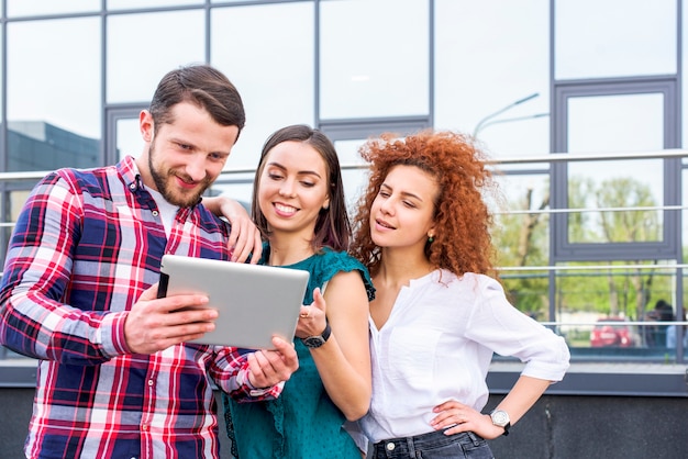 Foto amigos masculinos y femeninos jovenes felices que miran en la tableta digital que se coloca cerca del edificio de cristal