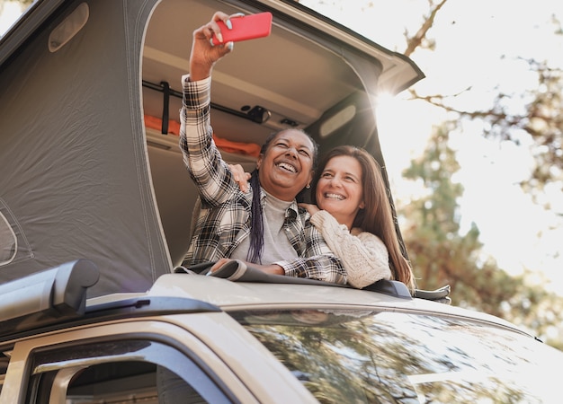 Amigos mayores multirraciales que toman un selfie en el techo de la caravana de la mini furgoneta usando el teléfono móvil