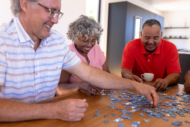 Foto amigos mayores multirraciales sonrientes arreglando piezas de rompecabezas sobre una mesa de madera en una casa de retiro. rompecabezas, resolución, juego, confusión, lluvia de ideas, inalterado, unión, apoyo, vida asistida.