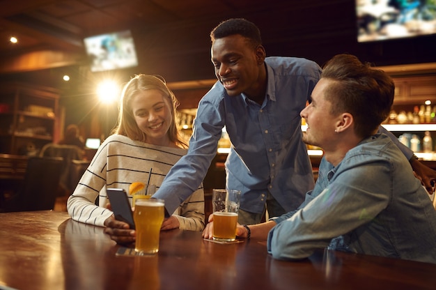 Los amigos miran fotos en el teléfono y se divierten en la mesa del bar. Grupo de personas se relajan en el pub, estilo de vida nocturno, amistad, celebración de eventos