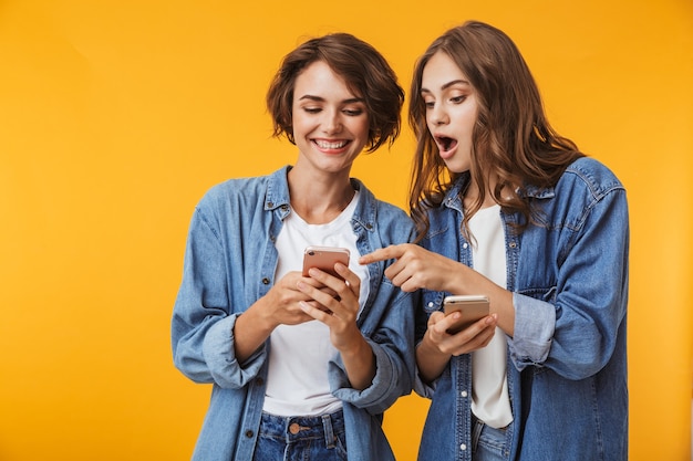 Amigos de mujeres jóvenes emocionales sorprendidos posando aislado sobre pared amarilla usando teléfonos móviles.