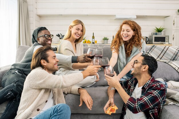 Amigos multiculturales jovenes alegres que tintinean con copas de vino tinto mientras hacen tostadas en el sofá en la fiesta de casa