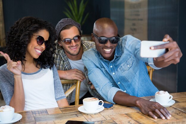 Amigos multiétnicos alegres con gafas de sol mientras toman selfie en cafetería