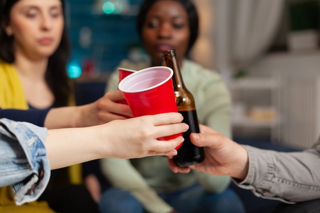 Amigos multiétnicos colgando sentados en el sofá charlando tintineando vasos de cerveza. Grupo de personas multirraciales que pasan tiempo juntos sentados en el sofá a altas horas de la noche en la sala de estar.