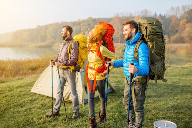Amigos multiétnicos en coloridas chaquetas de senderismo con mochilas, de pie cerca del camping en el césped verde