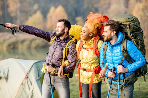 Amigos multiétnicos en coloridas chaquetas de senderismo con mochilas, de pie cerca del camping en el césped verde