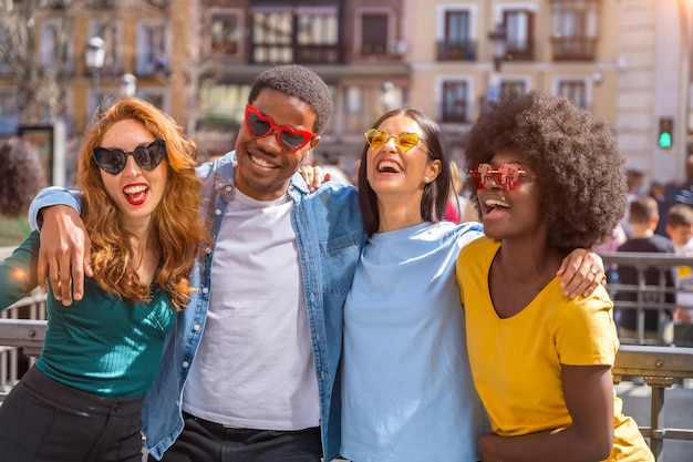 Amigos multiétnicos felizes usando óculos escuros de coração na rua da cidade ao pôr do sol rindo juntos ao ar livre