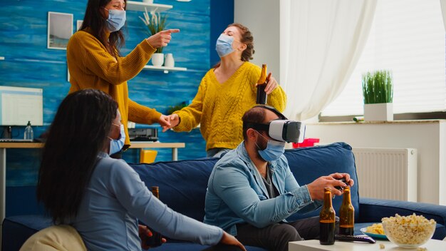 Amigos multiétnicos que disfrutan de la tecnología vr jugando videojuegos en la sala de estar con máscara para prevenir la infección por coronavirus manteniendo el distanciamiento social. Diversas personas divirtiéndose en la nueva fiesta normal.