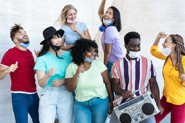 Foto amigos multirraciais dançando ao som de música com estéreo boombox ao ar livre com máscaras sob o queixo