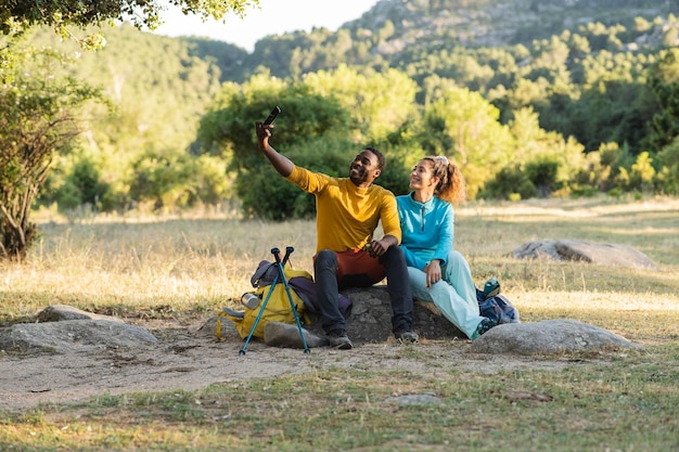 Amigos multirraciais na montanha sentados nas pedras descansando eles tiram uma selfie com smartphone mochileiros viajantes