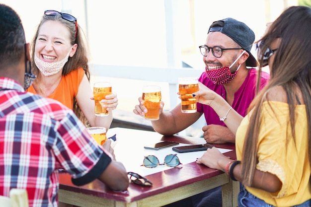 Amigos multirraciales animando con cerveza y sonriendo riendo entre ellos - Coronavirus / concepto de máscara facial