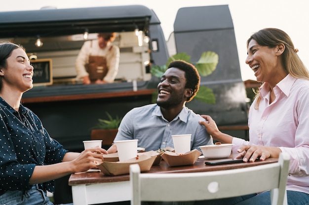 Amigos multirraciales comiendo en la mesa del camión de comida al aire libre - Concepto de verano y estilo de vida - Enfoque principal en el rostro de la mujer derecha