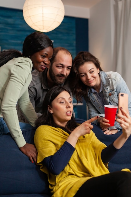 Amigos multirraciales pasando el rato tarde en la noche viendo videos de entretenimiento divertidos en el teléfono inteligente. Grupo de personas multirraciales que pasan tiempo juntos sentados en el sofá a altas horas de la noche en la sala de estar.