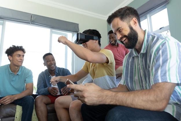 Foto amigos multirraciales riéndose y mirando a un hombre que usa un simulador de realidad virtual haciendo gestos en casa. copie el espacio, la tecnología, lo futurista, lo inalterado, la convivencia, el disfrute y las actividades de fin de semana.