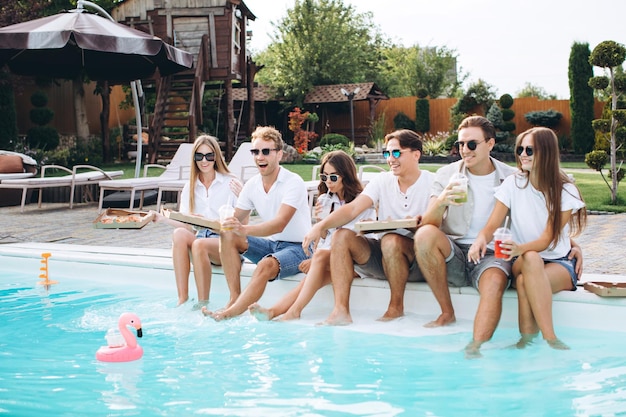 Amigos pasando el rato junto a la piscina. Comen pizza, beben cócteles y se divierten.