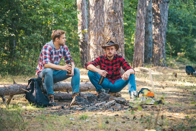 Amigos passam o fundo da natureza da floresta de fim de semana de lazer duas pessoas felizes sentadas ao redor da fogueira com cerveja amigos aproveitam o acampamento de fim de semana na floresta estilo de vida de verão