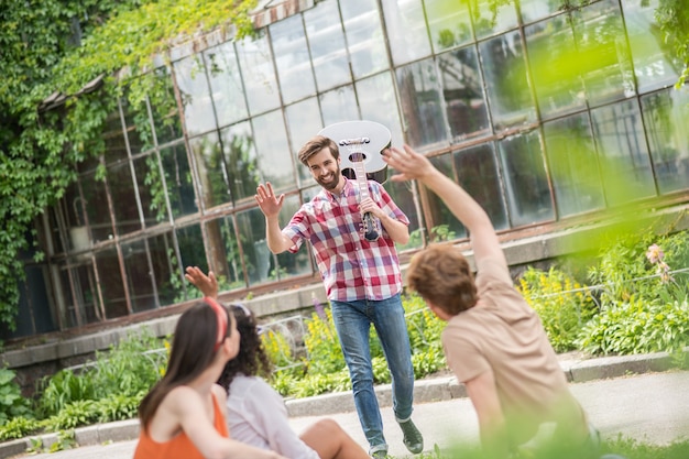 Amigos, picnic. Amigos sentados en una manta de espaldas a la cámara saludando a un conocido del joven guitarrista sonriente en el parque en una tarde agradable