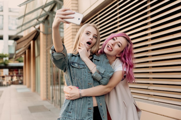 Amigos que toman selfie con un teléfono inteligente y hacen muecas y se divierten. Primer plano de chicas haciendo muecas y sonriendo para selfie. Dos amigas felices tomando fotos con su teléfono inteligente en la ciudad