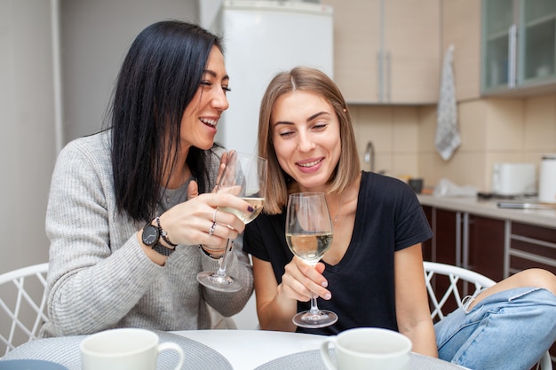 Amigos, reunião com vinho e bolo na cozinha de estilo moderno. as mulheres jovens sorriem e brincam com taças de vinho nas mãos