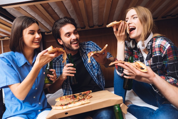 Los amigos se ríen, sostienen rebanadas de pizza y comen.