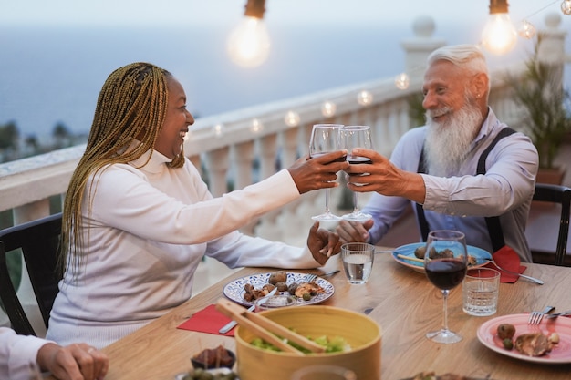 Amigos senior multirraciales animando con vino en el patio - Ancianos celebrando juntos mientras cenan en la terraza del restaurante