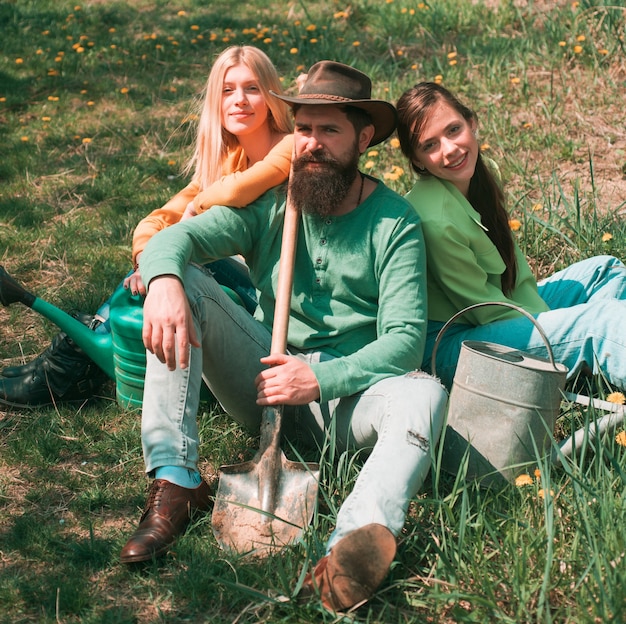 Amigos sentados en la hierba en el jardín de primavera. Retrato de agricultor caucásico con pala con dos amigas. Relajarse después del trabajo duro.