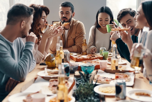 Amigos para siempre. Grupo de jóvenes en ropa casual comiendo pizza y sonriendo mientras tienen una cena en el interior