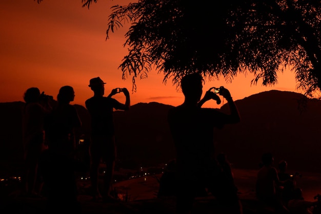 Foto amigos en silueta fotografiando mientras están de pie en la playa durante la puesta de sol