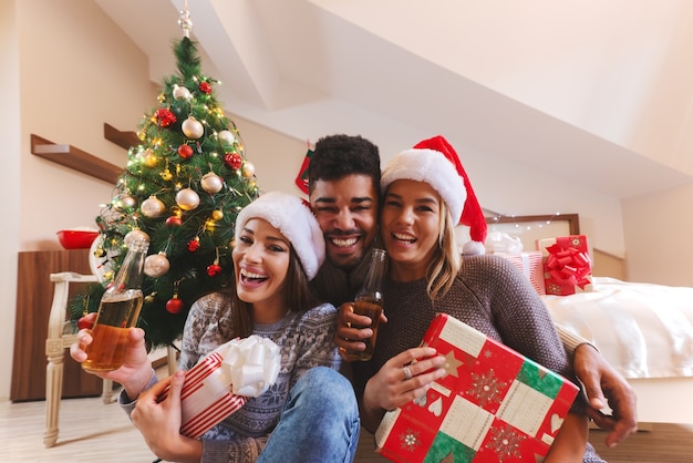 Amigos sonrientes posando con regalos y botellas de cerveza en las manos en el dormitorio. Concepto de vacaciones de Navidad.