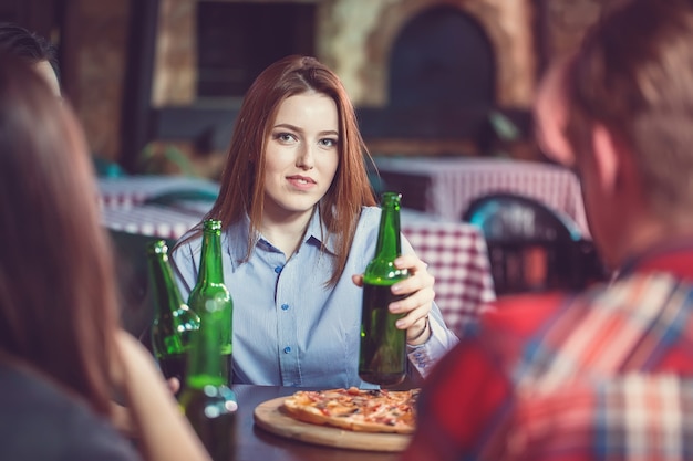 Foto amigos tomando una copa en un bar, están sentados en una mesa de madera con cervezas y pizza. centrarse en una hermosa niña tocando su botella.