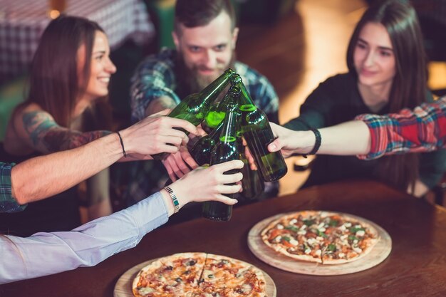 Amigos tomando una copa en un bar, están sentados en una mesa de madera con cervezas y pizza.