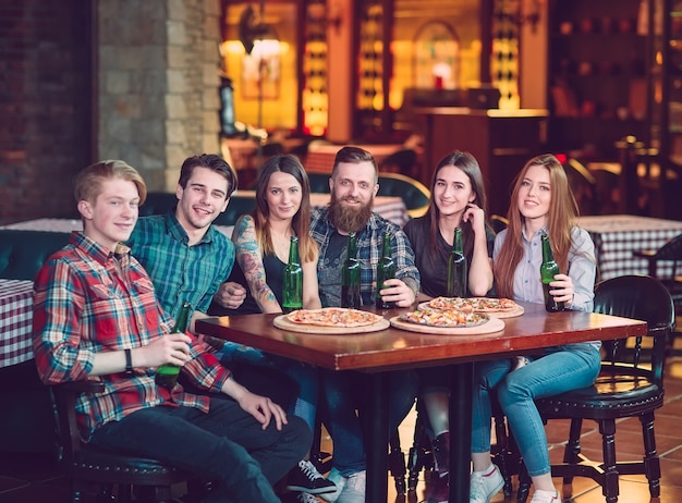 Amigos tomando una copa en un bar, están sentados en una mesa de madera con cervezas y pizza.