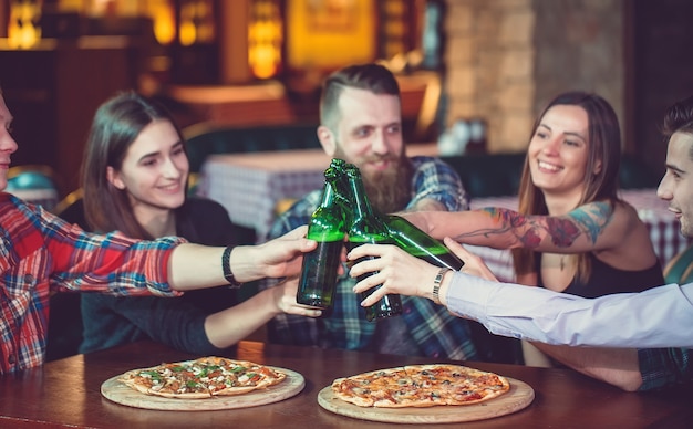 Amigos tomando una copa en un bar, están sentados en una mesa de madera con cervezas y pizza.