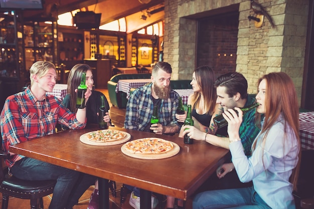 Amigos tomando una copa en un bar, están sentados en una mesa de madera con cervezas y pizza.