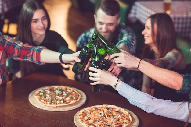 Amigos tomando una copa en un bar, están sentados en una mesa de madera con cervezas y pizza.