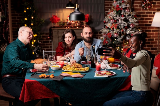 Amigos tomando fotografías en víspera de Navidad, creando hermosos recuerdos de festividad en casa. Diversas personas divirtiéndose con vino y comida, disfrutando de fotografías para recuerdos en la mesa.