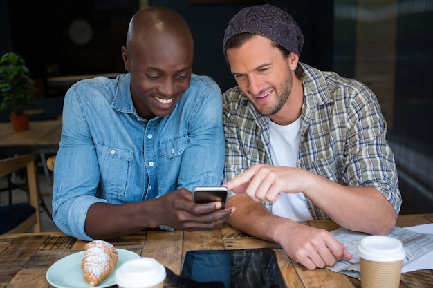 Amigos varones felices a través de teléfono móvil en la mesa en la casa de café