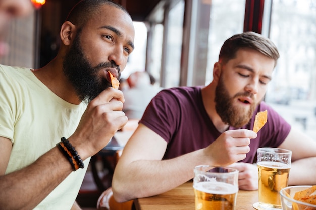 Amigos varones guapos bebiendo cerveza y comiendo patatas fritas en el pub